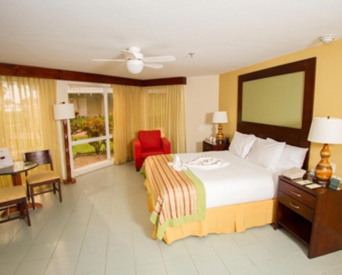 Fiesta Resort All Inclusive Central Pacific - Costa Rica El Roble Room photo The photo shows a hotel room that includes a queen-sized bed with white linens and a colorful throw blanket. There are two bedside tables with lamps on either side of the bed. A small red armchair is placed in one corner of the room, adding a pop of 
