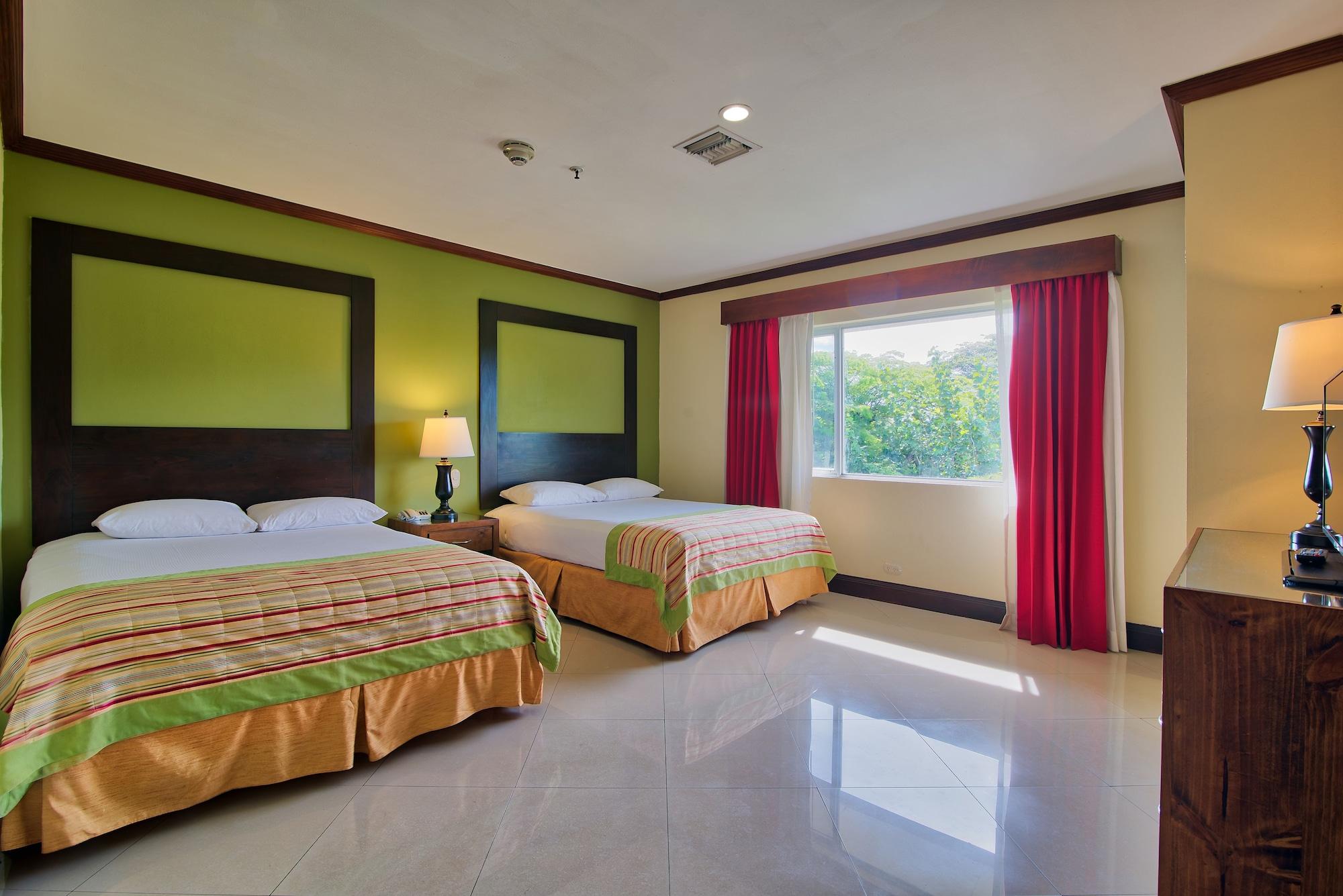 Fiesta Resort All Inclusive Central Pacific - Costa Rica El Roble Exterior photo The photo shows a hotel room featuring two beds with colorful bedding. One bed is positioned against a green wall with a decorative headboard. There is a bedside lamp next to one of the beds. The room has large windows with red curtains, allowing nat
