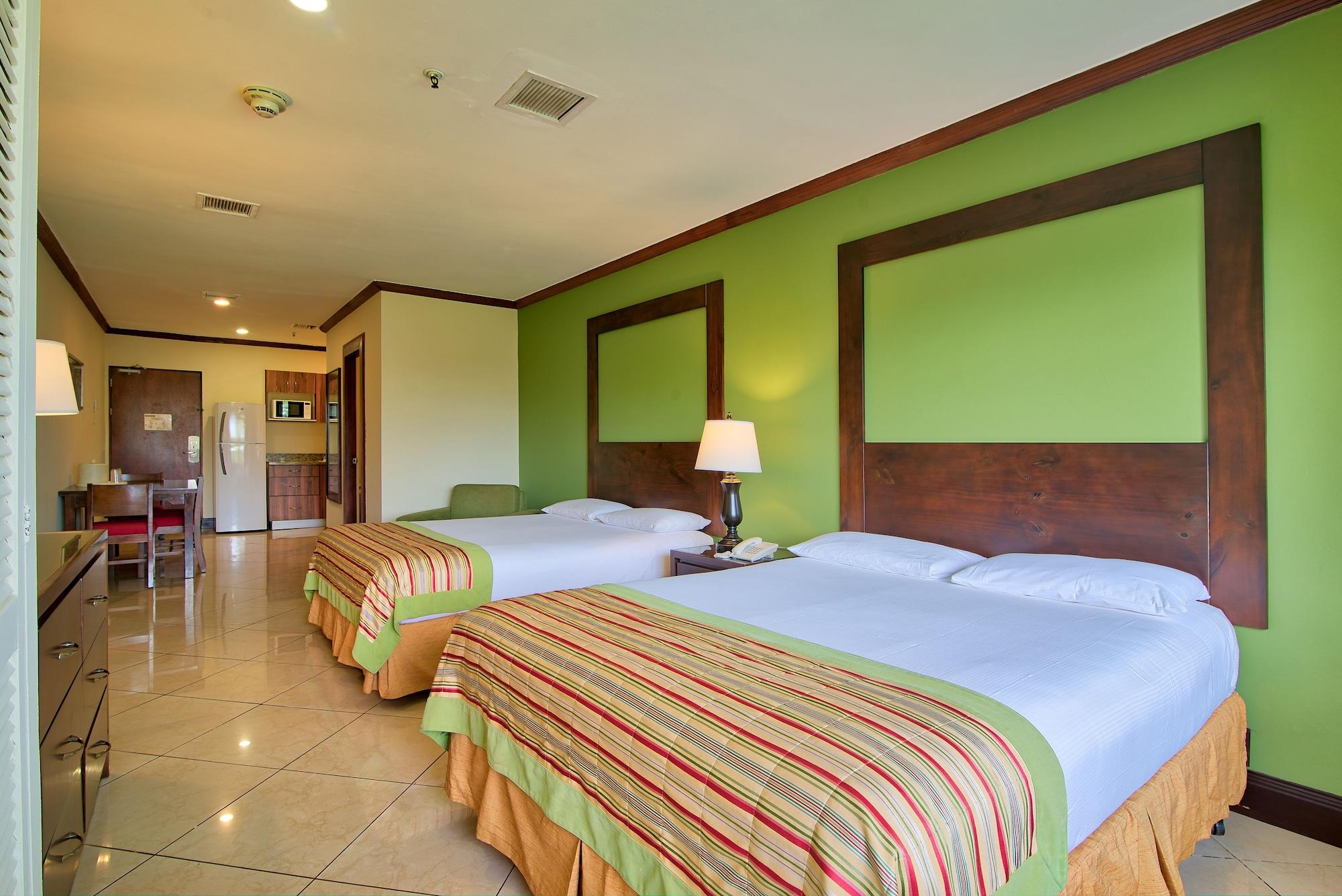 Fiesta Resort All Inclusive Central Pacific - Costa Rica El Roble Exterior photo The photo shows a hotel room featuring two beds with colorful striped bedspreads. The walls are painted a bright green, and there is a wooden headboard for each bed. A bedside lamp is placed between the beds. In the background, a kitchenette area is 