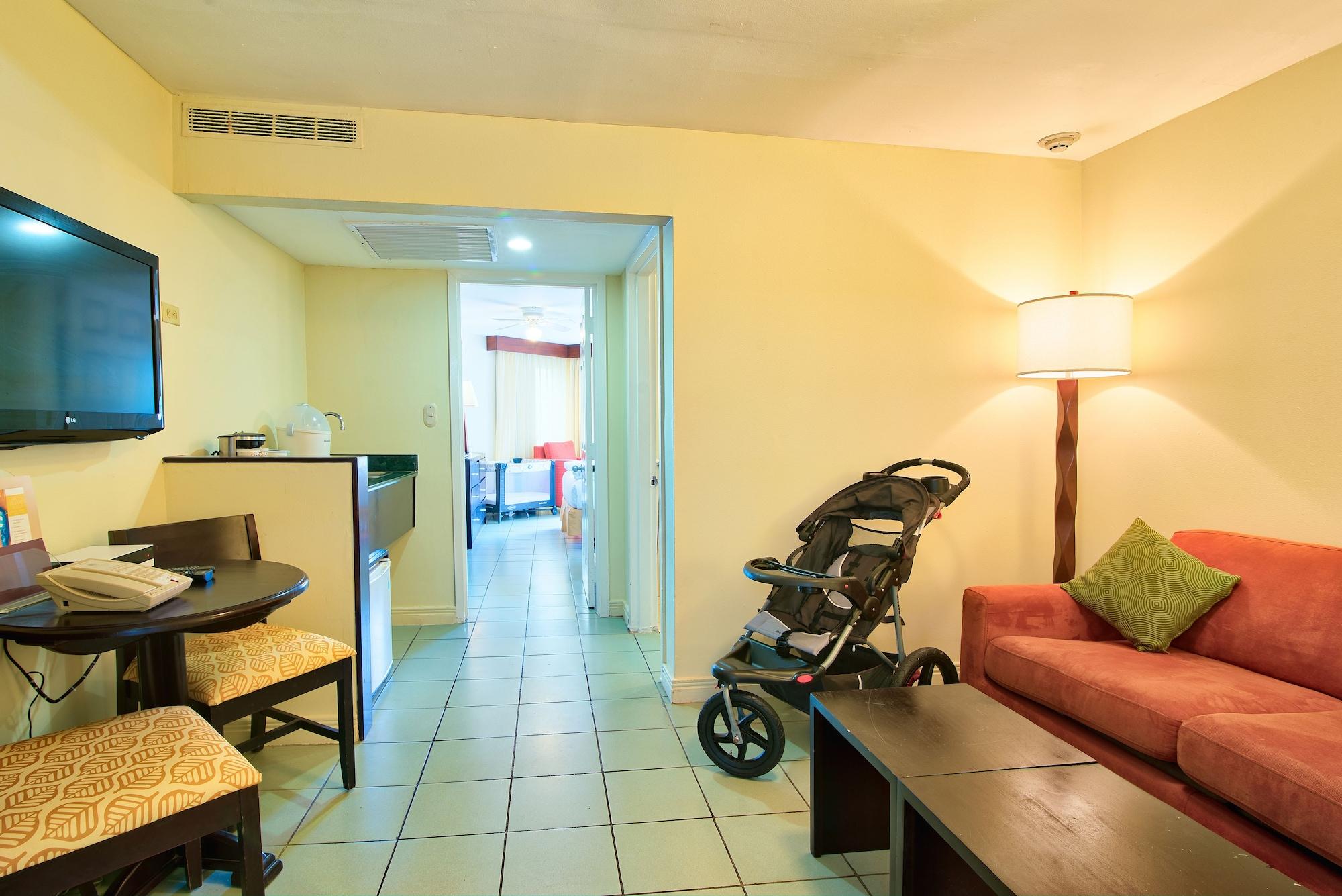 Fiesta Resort All Inclusive Central Pacific - Costa Rica El Roble Exterior photo The photo displays a small living area of an accommodation. There is a red sofa with a green cushion, a couple of dark wooden coffee tables, and a small dining table with two chairs. A wall-mounted flat-screen TV is visible, along with a refrigerator