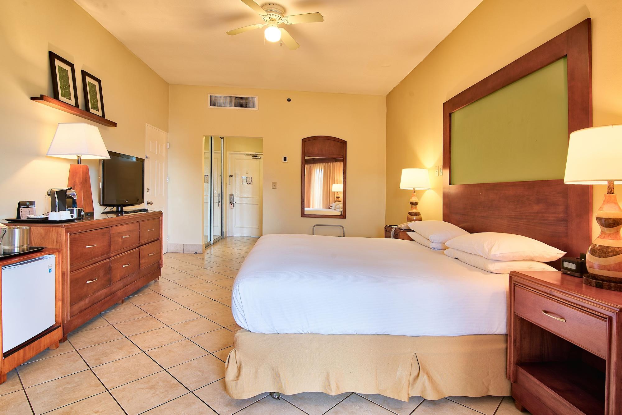 Fiesta Resort All Inclusive Central Pacific - Costa Rica El Roble Exterior photo The photo shows a hotel room. It features a comfortable bed with white linens and a light-colored bed skirt. To the side, there is a wooden nightstand with a lamp. There is a large wooden dresser with a television on top, and a coffee maker is also p