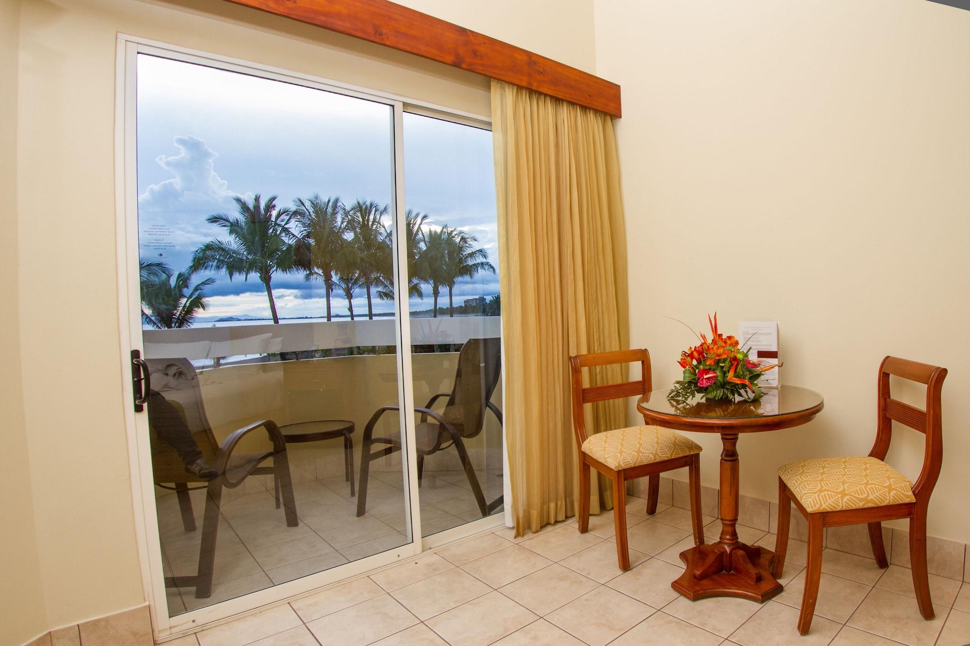 Fiesta Resort All Inclusive Central Pacific - Costa Rica El Roble Exterior photo The photo shows a room with a large glass sliding door that opens to a balcony or patio area. There are two outdoor chairs on the balcony, allowing for a view of the outdoors, including palm trees in the background. Inside the room, there is a small 