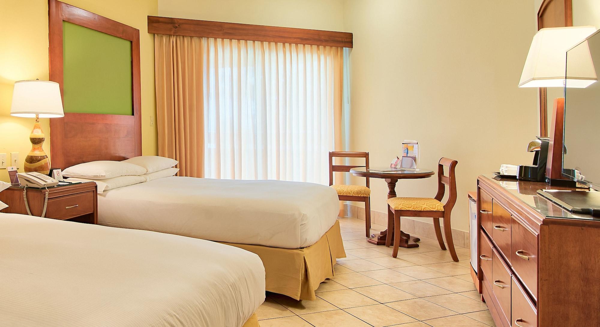 Fiesta Resort All Inclusive Central Pacific - Costa Rica El Roble Exterior photo The photo shows a hotel room featuring two beds with light-colored linens. There is a small round table with two chairs positioned near a large window that has light curtains. The walls are a soft yellow color, and there is a green accent wall behind