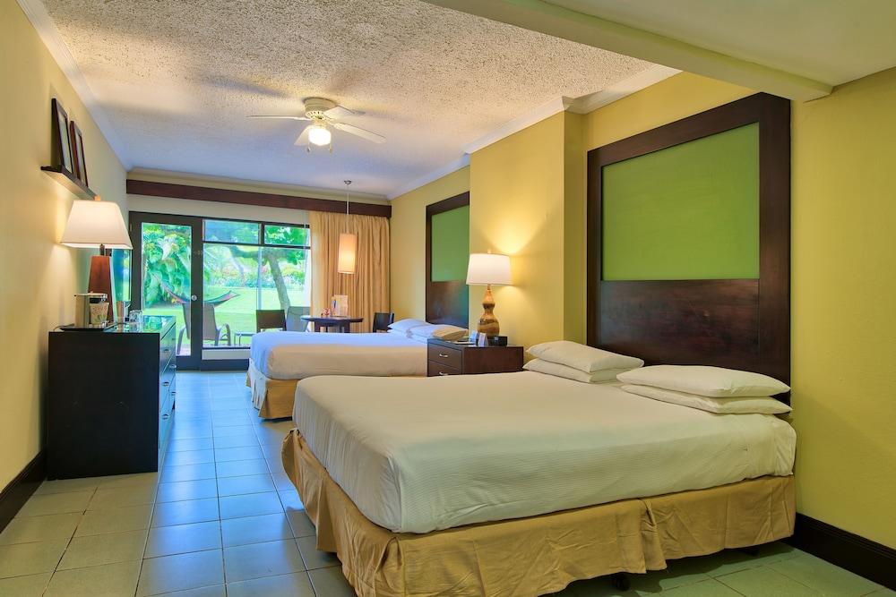Fiesta Resort All Inclusive Central Pacific - Costa Rica El Roble Exterior photo The photo shows a hotel room featuring two beds with white linens and light brown bed skirts. The walls are painted in a warm yellow tone, with green accents behind the headboards. There are lamps on bedside tables next to each bed. A dresser is visi
