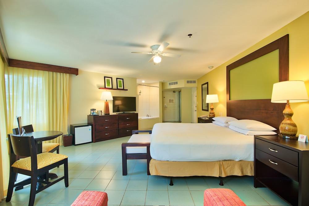 Fiesta Resort All Inclusive Central Pacific - Costa Rica El Roble Exterior photo The photo shows a hotel room featuring a spacious layout. There is a large bed with white bedding positioned against a wall with a decorative headboard. On either side of the bed, there are bedside lamps. In the room, there's a wooden dresser with a 