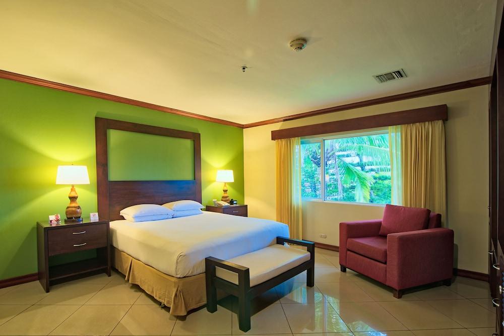 Fiesta Resort All Inclusive Central Pacific - Costa Rica El Roble Exterior photo The photo shows a hotel room with a modern design. It features a double bed with white bedding and a wooden headboard, flanked by bedside tables with lamps. The walls are painted a vibrant green, creating a lively atmosphere. There's a window with li
