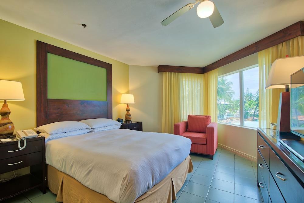 Fiesta Resort All Inclusive Central Pacific - Costa Rica El Roble Exterior photo The photo shows a hotel room featuring a queen-sized bed with white bedding. There is a wooden headboard behind the bed, and on either side, there are bedside lamps. To the right of the bed, there's a small red armchair and a window with yellow curta