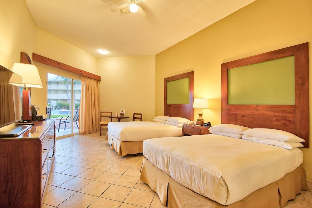 Fiesta Resort All Inclusive Central Pacific - Costa Rica El Roble Exterior photo The photo shows a hotel room with two queen-sized beds. The walls are painted in a warm yellow hue, and each bed has a light-colored comforter. There are wooden headboards with green panels above the beds. A small table and two chairs are visible nea