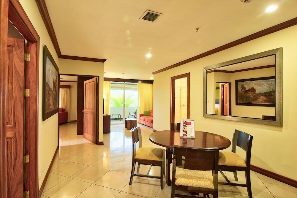 Fiesta Resort All Inclusive Central Pacific - Costa Rica El Roble Exterior photo The photo shows a spacious and well-lit interior of a hotel room or suite. There is a small dining table with four chairs in the center, set on a tiled floor. The walls are painted in a light color, possibly cream, with wooden accents around the door