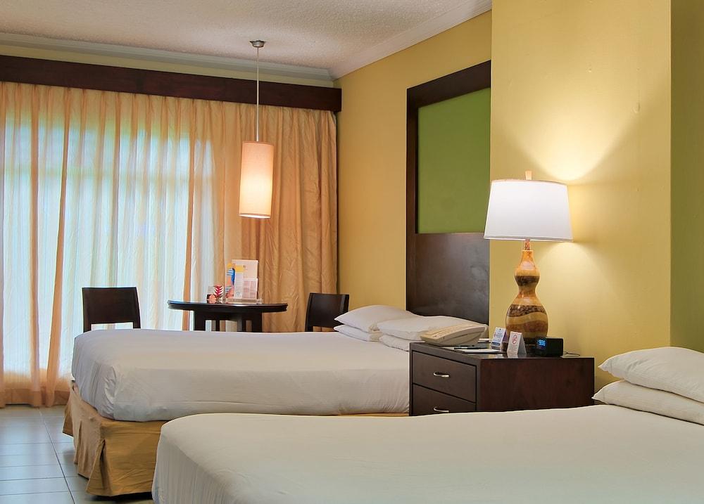 Fiesta Resort All Inclusive Central Pacific - Costa Rica El Roble Exterior photo The photo shows a hotel room featuring two beds with white linens. The room has light yellow walls, a large window covered by sheer curtains, and a lampshade hanging from the ceiling. There is a small round table with two chairs near the window, and 
