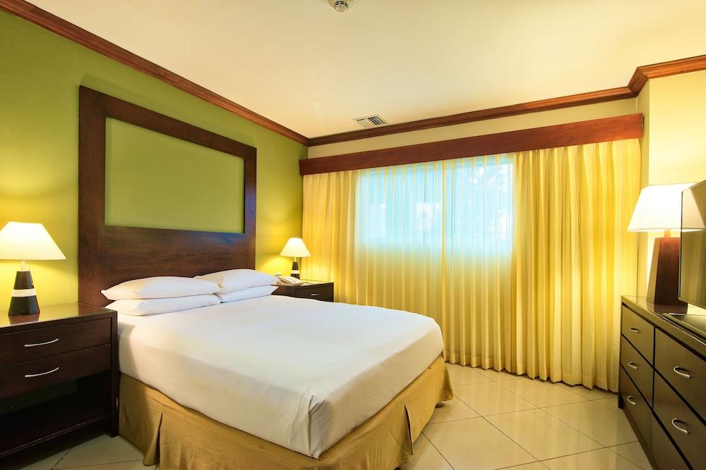 Fiesta Resort All Inclusive Central Pacific - Costa Rica El Roble Exterior photo The photo shows a simple and inviting hotel room. It features a double bed with white bedding, placed against a light green wall that has a dark wooden headboard. On either side of the bed, there are nightstands with lamps. A large window covered wit