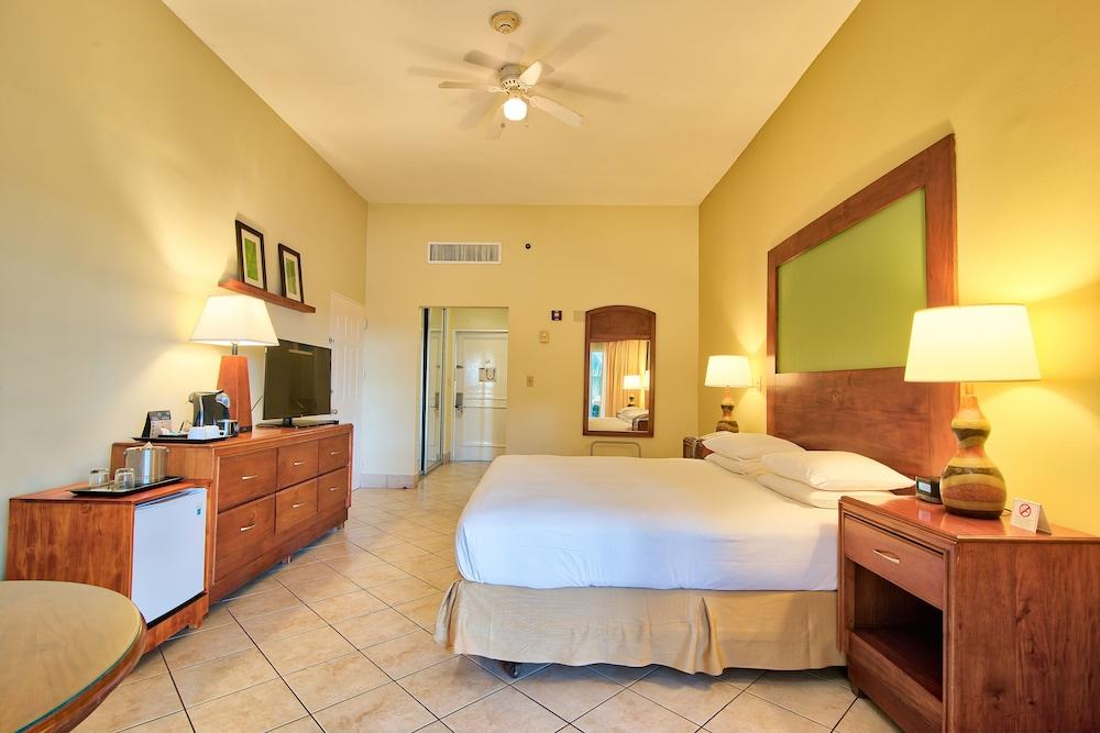 Fiesta Resort All Inclusive Central Pacific - Costa Rica El Roble Exterior photo The photo shows an interior of a hotel room. The room features a double bed with a light-colored bedspread. There are wooden furniture pieces, including a bedside table with lamps and a dresser with a television on top. A small fridge is located besi