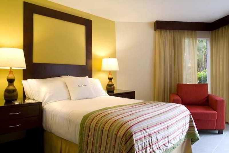 Fiesta Resort All Inclusive Central Pacific - Costa Rica El Roble Exterior photo The photo shows a hotel room featuring a neatly made bed with white linens and a colorful striped blanket at the foot. There are two bedside lamps on either side of the bed, placed on dark wooden nightstands. The wall behind the bed is painted a warm