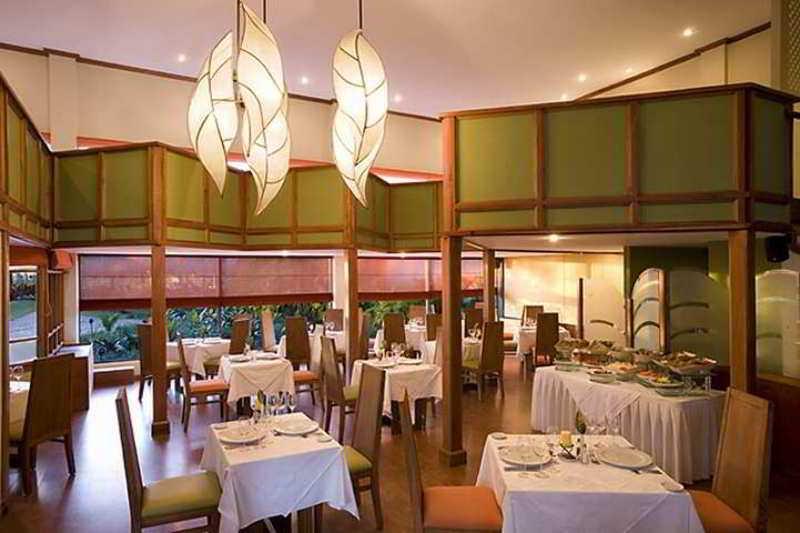 Fiesta Resort All Inclusive Central Pacific - Costa Rica El Roble Restaurant photo The photo depicts an elegantly designed restaurant interior. The space features a combination of wooden elements and green accents, creating a warm and inviting atmosphere. 

There are multiple dining tables covered with white tablecloths, set with p