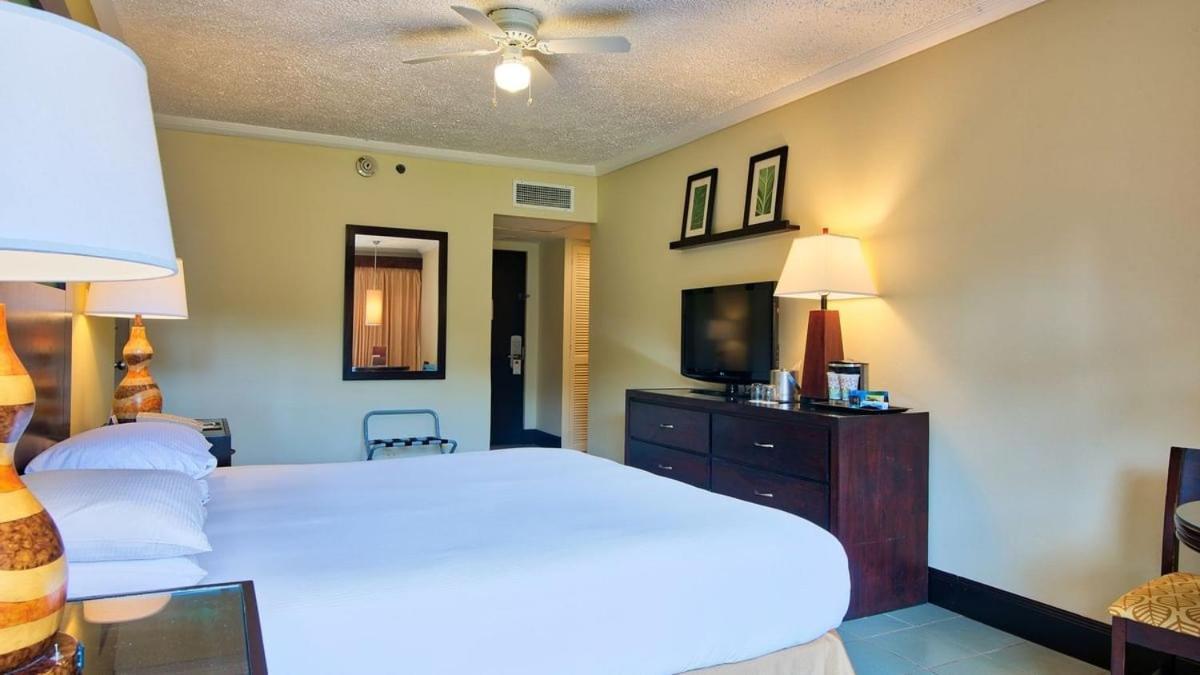 Fiesta Resort All Inclusive Central Pacific - Costa Rica El Roble Exterior photo The photo shows a hotel room featuring a comfortable bed with white linens. To the right of the bed, there is a wooden dresser with a television on top, and a lamp beside it. The wall has framed artwork or photographs. There is a mirror on the wall, 
