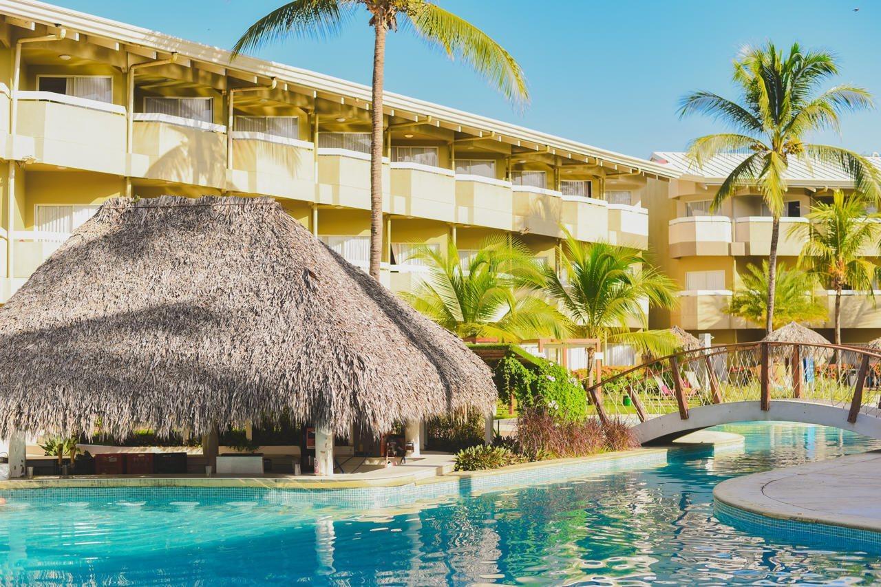 Fiesta Resort All Inclusive Central Pacific - Costa Rica El Roble Exterior photo The photo shows a tropical resort setting. In the foreground, there is a swimming pool with a clear blue surface. Nearby, a thatched-roof hut provides a shaded area, likely for relaxation. Lush palm trees surround the area, enhancing the tropical atm