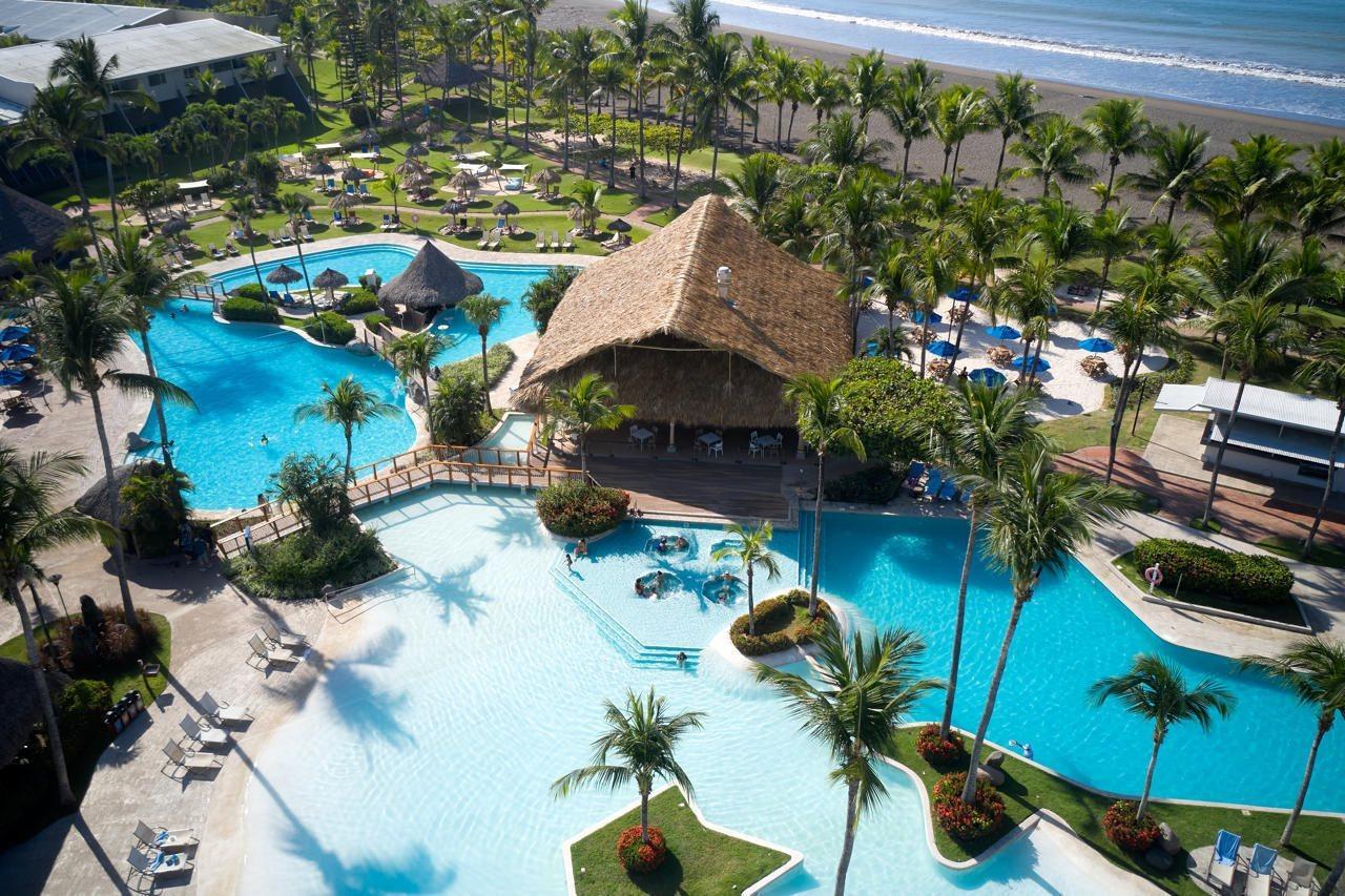 Fiesta Resort All Inclusive Central Pacific - Costa Rica El Roble Exterior photo The photo shows a scenic view of a resort area featuring multiple swimming pools, surrounded by lush greenery and palm trees. In the center, there is a tiki-style hut that likely serves as a bar or lounge area. The pools are shaped irregularly, creat