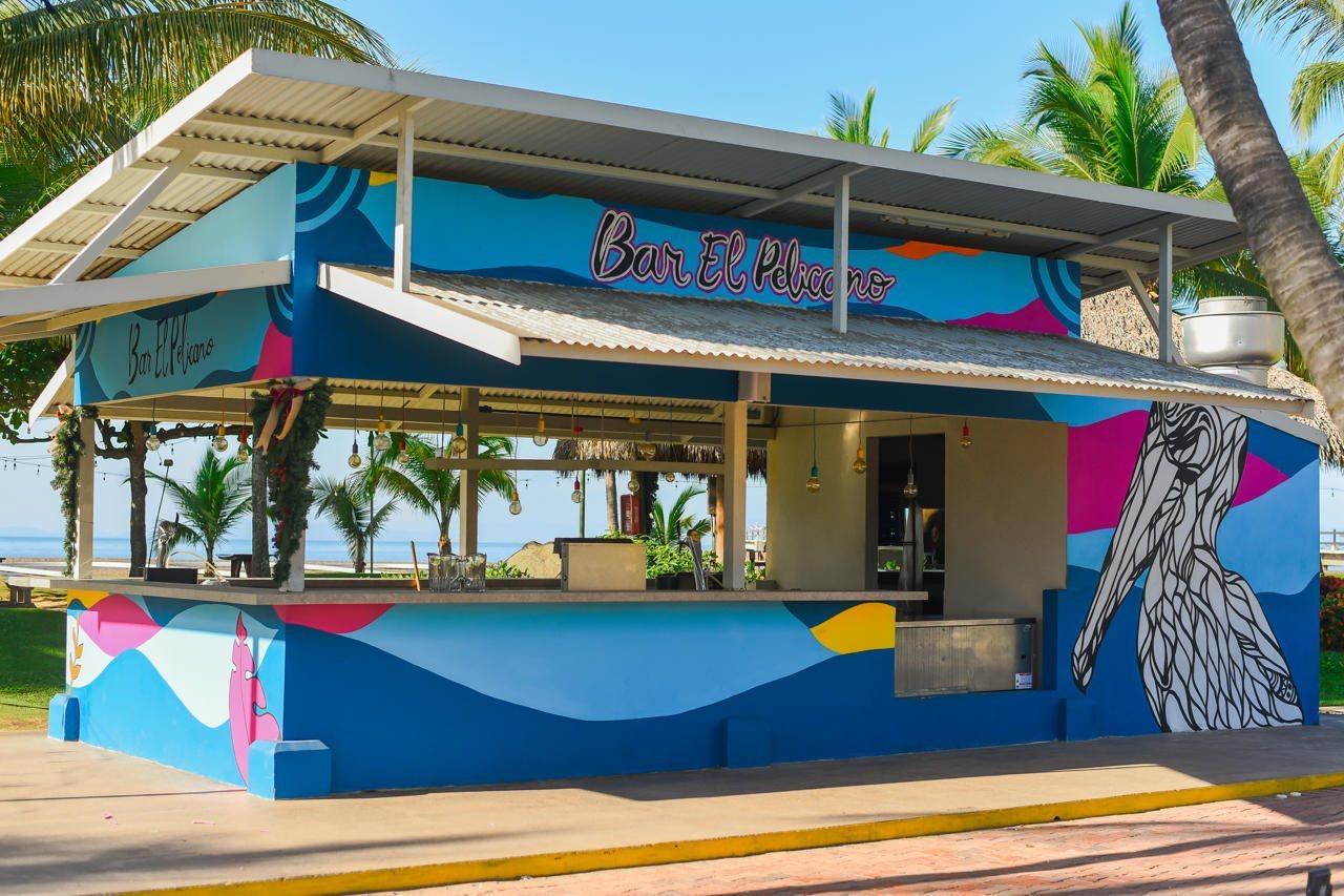 Fiesta Resort All Inclusive Central Pacific - Costa Rica El Roble Exterior photo The photo shows a vibrant bar structure named "Bar El Placer." It features colorful murals with waves and a large depiction of a bird, likely a pelican. The bar has an open design, allowing for a view of the surrounding palm trees and presumably a be