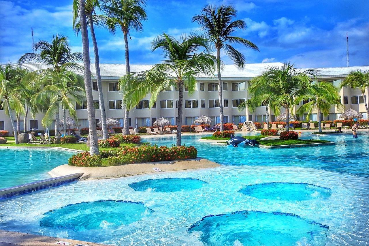 Fiesta Resort All Inclusive Central Pacific - Costa Rica El Roble Exterior photo The photo shows a resort swimming pool area surrounded by lush palm trees and colorful landscaping. The pool features distinct circular areas, possibly hot tubs or shallow spots, within the larger pool structure. In the background, there is a white b