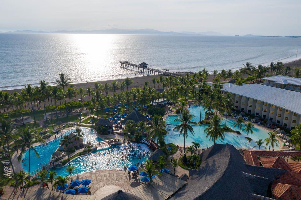 Fiesta Resort All Inclusive Central Pacific - Costa Rica El Roble Exterior photo The photo shows a beautiful coastal scene featuring a resort with multiple swimming pools surrounded by palm trees. There is a sandy beach leading to the ocean, with gentle waves and a distant pier extending into the water. The sunlight reflects on t