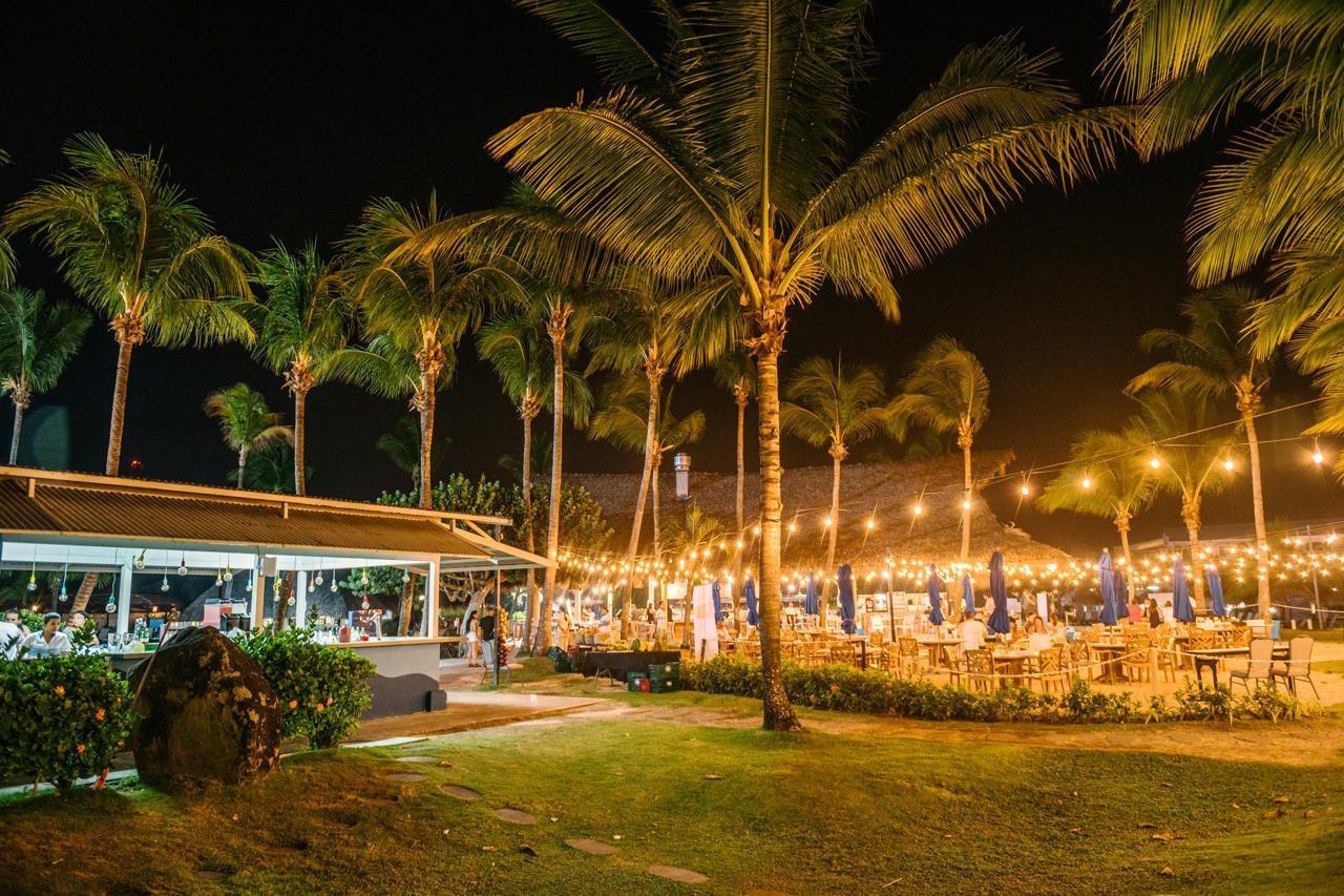 Fiesta Resort All Inclusive Central Pacific - Costa Rica El Roble Exterior photo The photo depicts a tropical outdoor dining area at night. There are numerous palm trees swaying gently, illuminated by festive string lights. The setting features a well-lit restaurant or bar area to the left, with tables set up for patrons. In the 