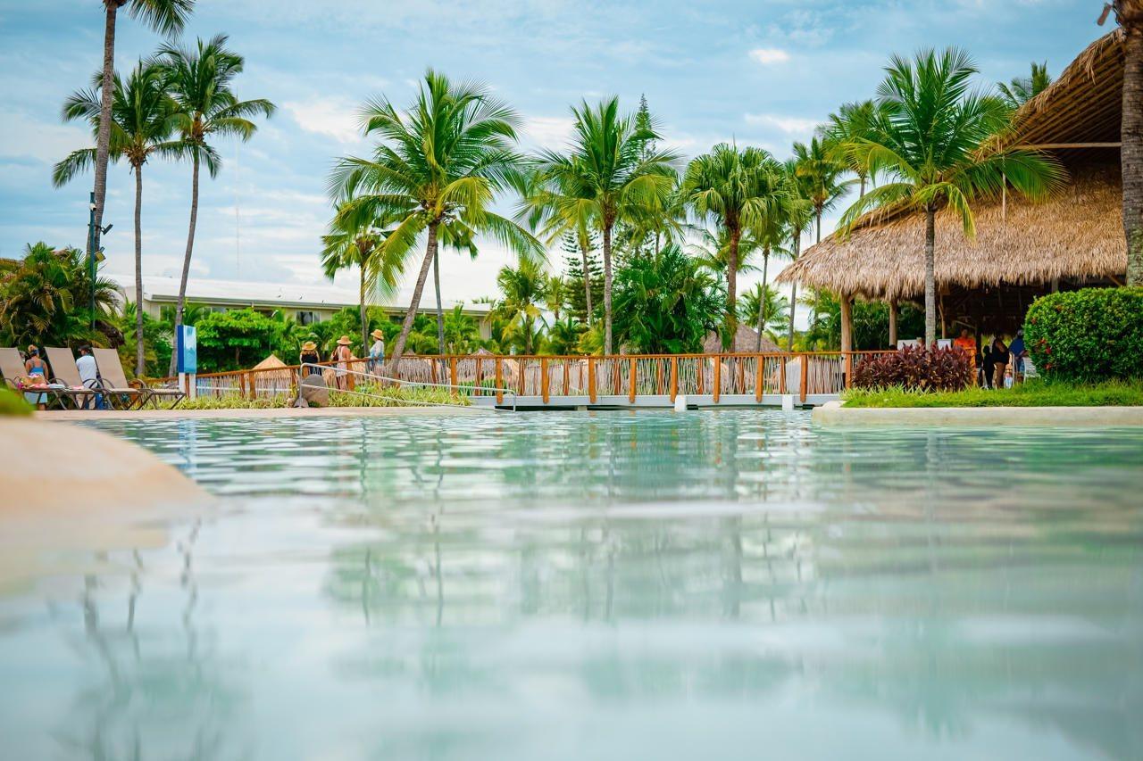 Fiesta Resort All Inclusive Central Pacific - Costa Rica El Roble Exterior photo The photo shows a serene pool area surrounded by palm trees and tropical vegetation. The water in the pool is calm and reflects the sky above, creating a tranquil atmosphere. In the background, there is a thatched-roof structure, possibly a cabana or