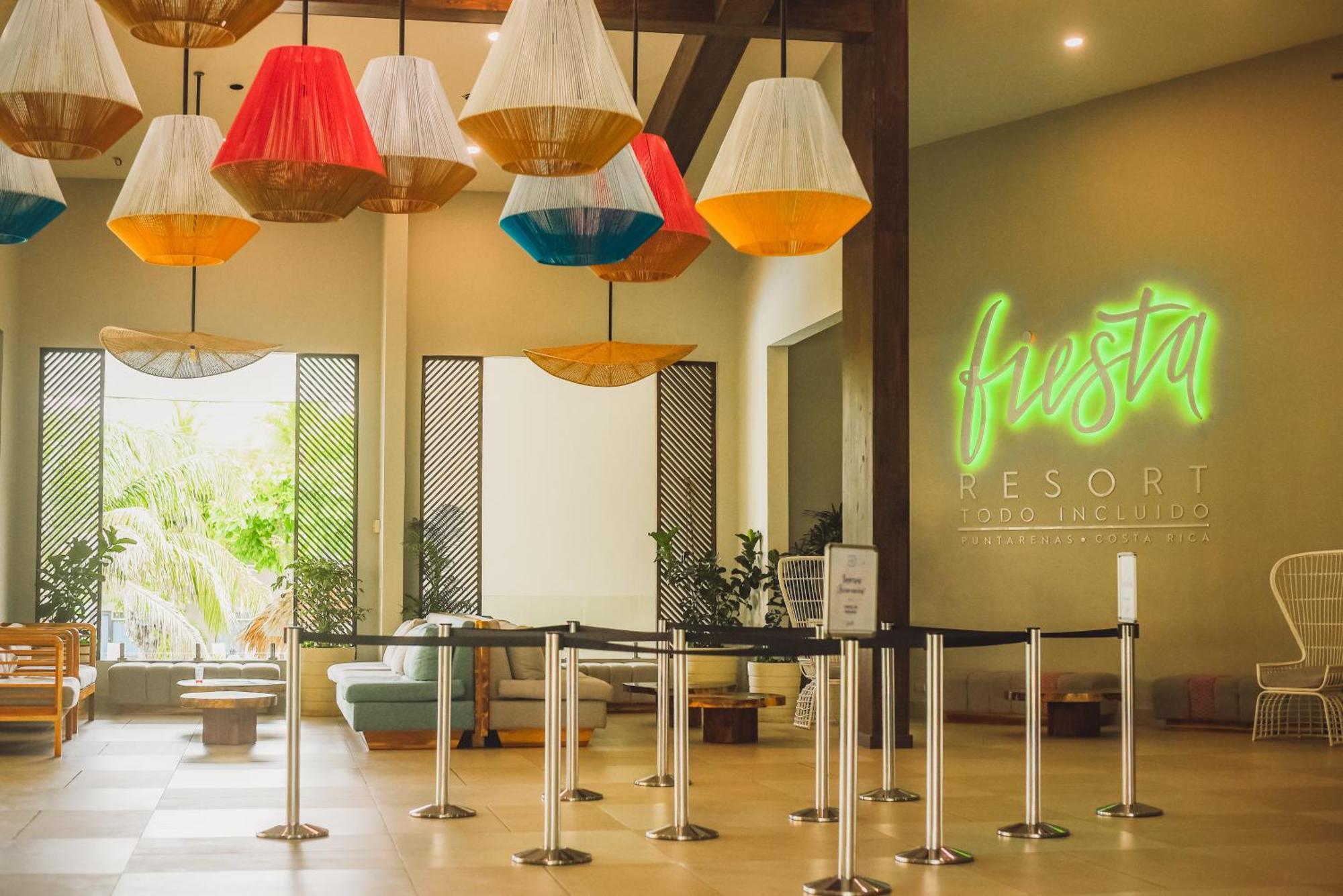 Fiesta Resort All Inclusive Central Pacific - Costa Rica El Roble Exterior photo The photo depicts the interior of a resort lobby. The space features colorful, large pendant lights hanging from the ceiling, adding a vibrant touch to the atmosphere. On one wall, there is a glowing neon sign that reads "Fiesta Resort." The lobby al