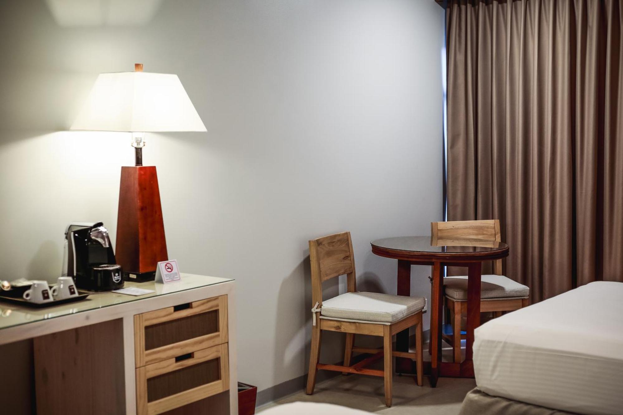 Fiesta Resort All Inclusive Central Pacific - Costa Rica El Roble Exterior photo The photo shows a cozy room interior, featuring a small table with two wooden chairs. To the left, there is a lamp on a wooden stand and a coffee machine on a cabinet with drawers. The wall is painted a light color, and the window is adorned with bei