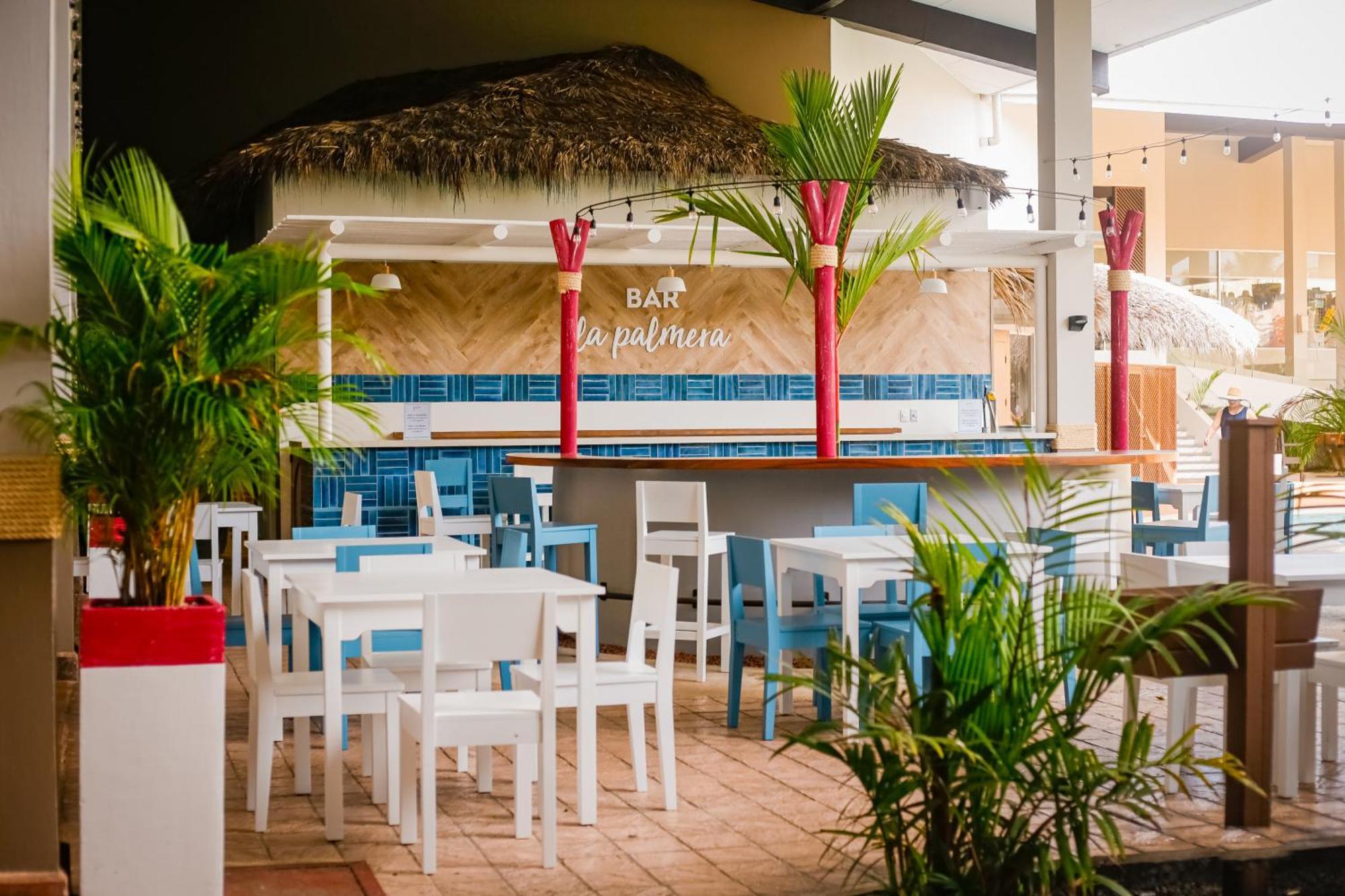 Fiesta Resort All Inclusive Central Pacific - Costa Rica El Roble Exterior photo The photo shows a tropical bar setting with a thatched roof and a vibrant atmosphere. In the foreground, there are several white wooden tables and chairs arranged neatly. The bar area itself is adorned with colorful decorations, including red pillars