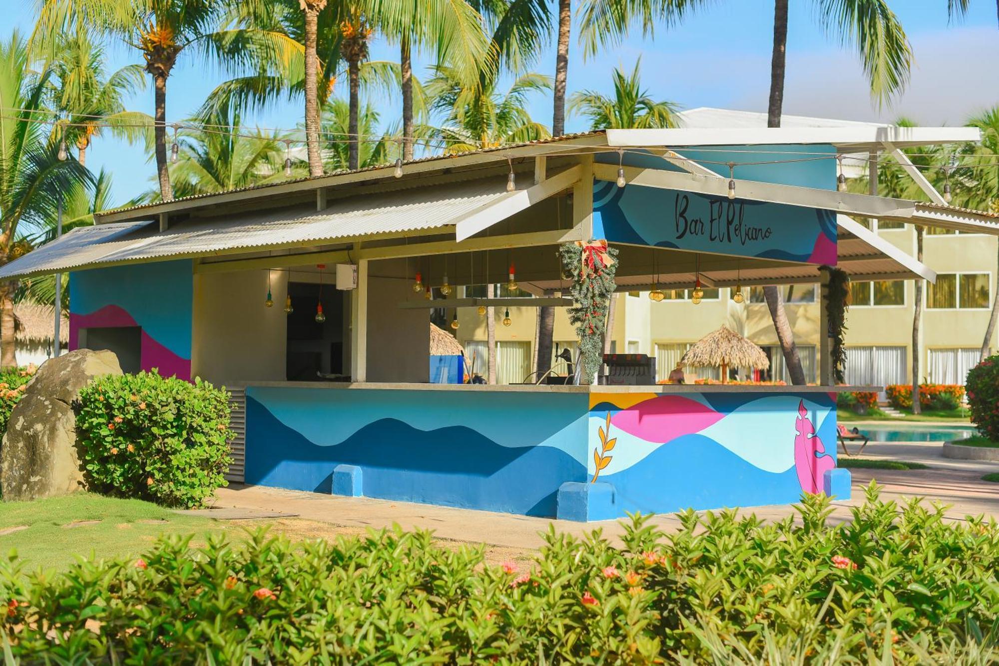 Fiesta Resort All Inclusive Central Pacific - Costa Rica El Roble Exterior photo The photo shows a colorful outdoor bar or kiosk situated in a tropical setting. It features a roof and vibrant painted designs in shades of blue, pink, and purple. Surrounding the bar are lush green bushes and palm trees, indicating a warm, sunny env