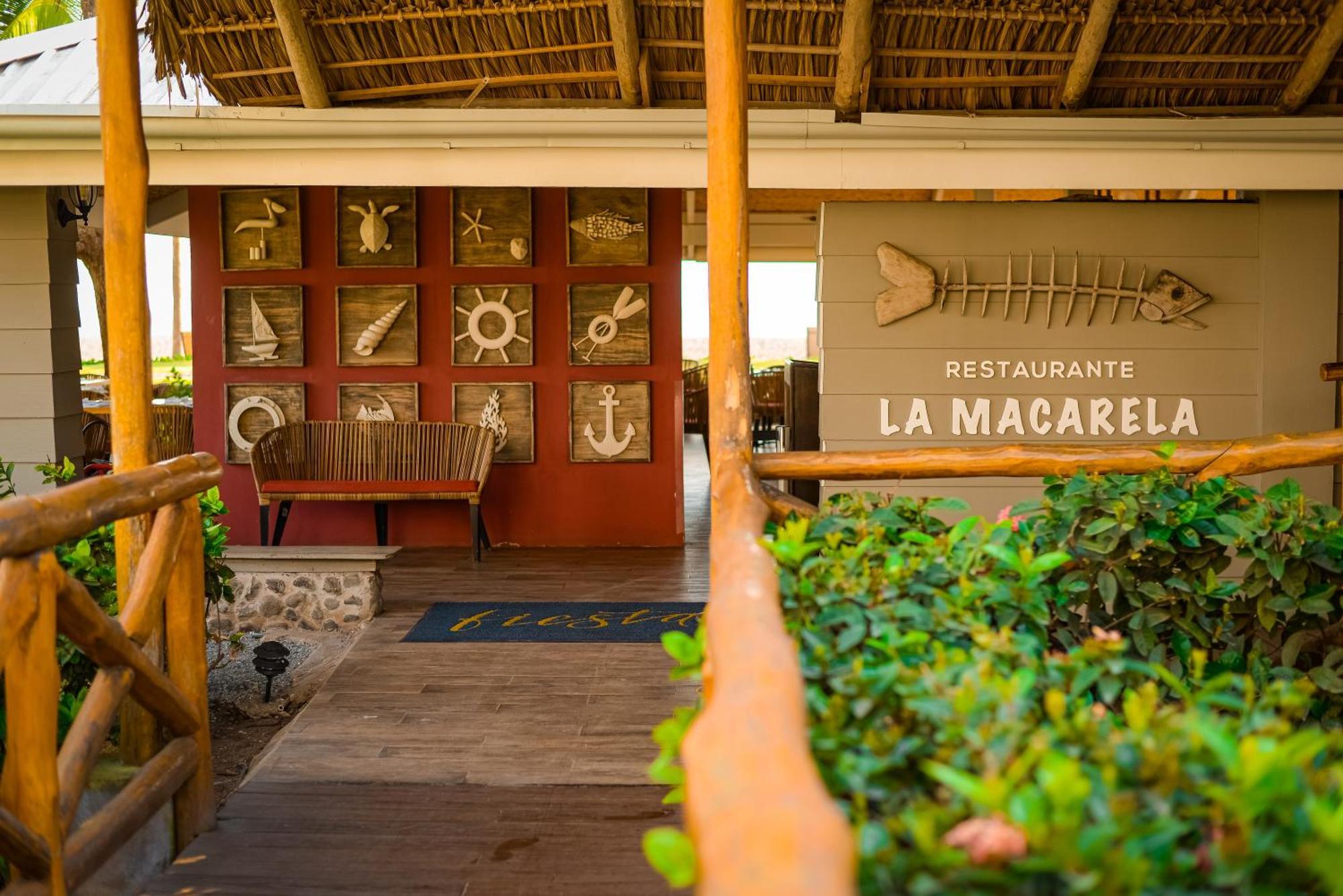 Fiesta Resort All Inclusive Central Pacific - Costa Rica El Roble Exterior photo The photo shows the entrance to a restaurant named "La Macarela." The exterior features a wooden pathway leading up to the entrance, with a bench on one side. The wall beside the entrance displays a series of decorative elements, including various ma