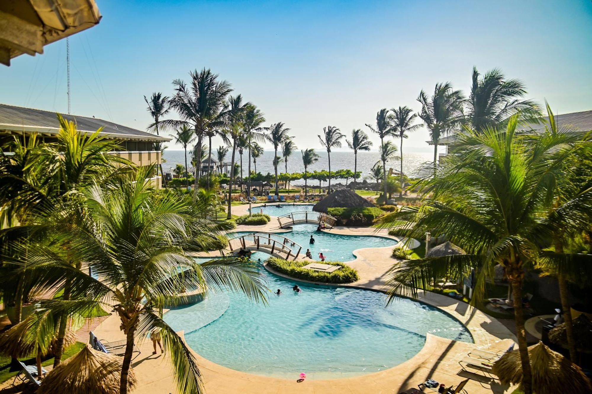 Fiesta Resort All Inclusive Central Pacific - Costa Rica El Roble Exterior photo The photo depicts a vibrant tropical scene featuring a large swimming pool surrounded by palm trees. The pool area has a curved shape and is situated in a sunny environment, suggesting a warm climate. There are several people enjoying the water, and 