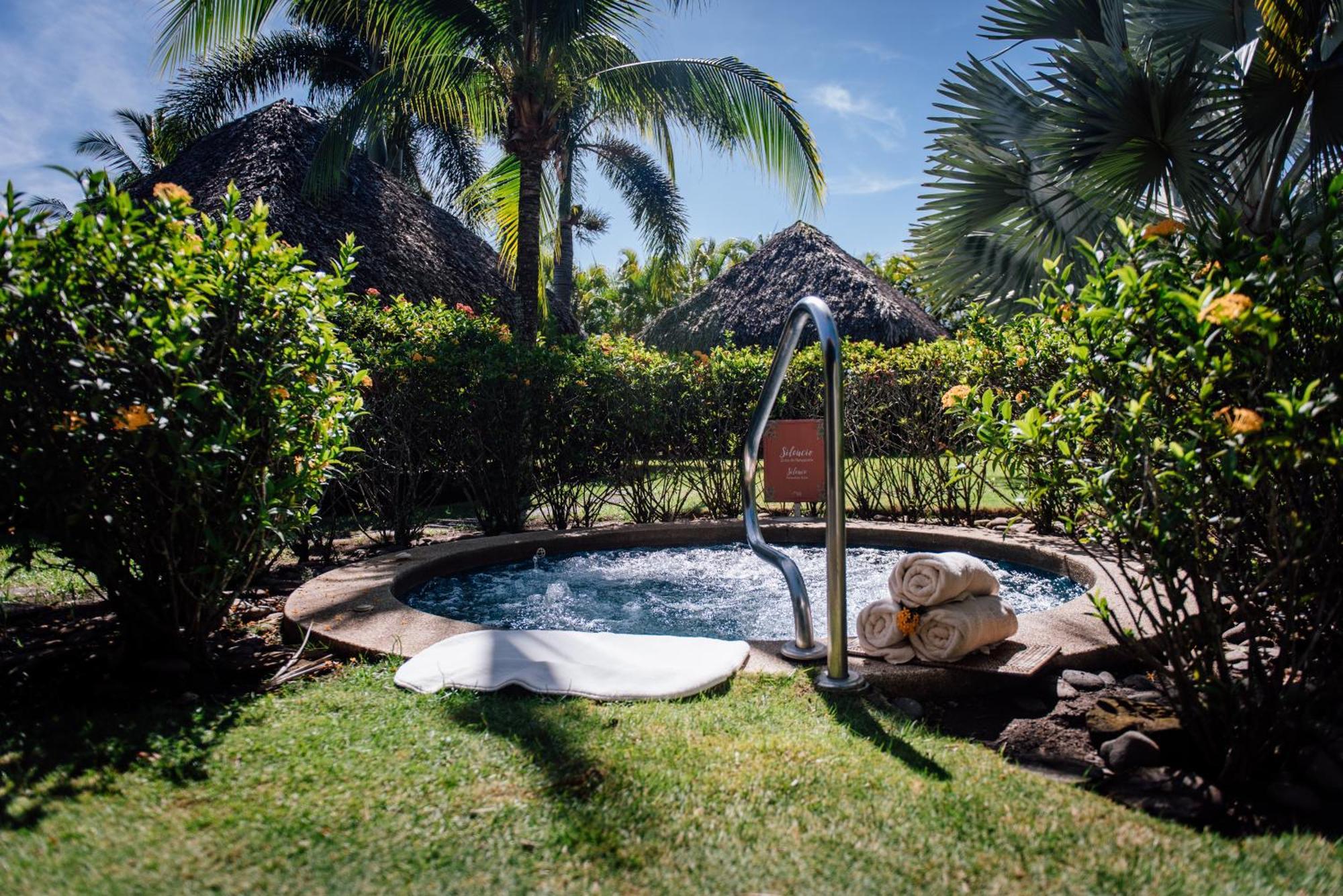 Fiesta Resort All Inclusive Central Pacific - Costa Rica El Roble Exterior photo The photo features a relaxing outdoor setting with a hot tub surrounded by lush greenery. There are palm trees and ornamental bushes enclosing the area, creating a serene atmosphere. Next to the hot tub, neatly stacked towels are placed on a white bl