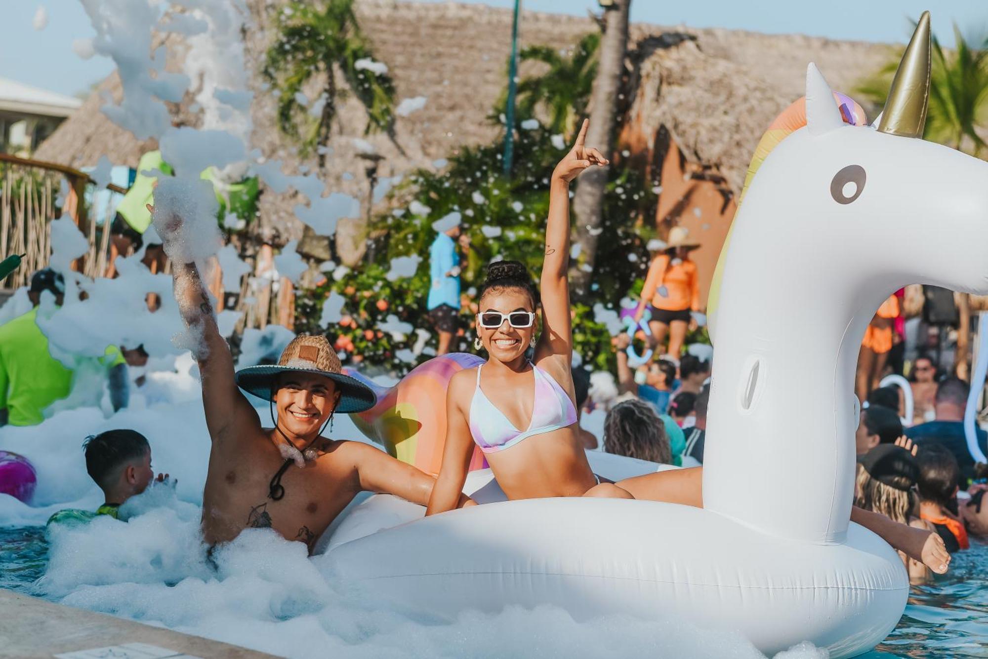 Fiesta Resort All Inclusive Central Pacific - Costa Rica El Roble Exterior photo The photo shows a lively pool scene filled with foam. In the foreground, a young woman is sitting on a large inflatable unicorn, wearing a bikini, sunglasses, and smiling while giving a peace sign. Next to her, a man is sitting in the foam, wearing a
