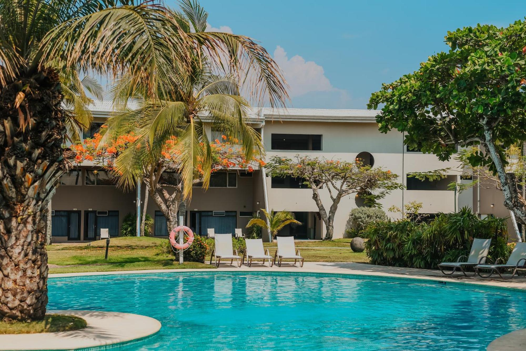 Fiesta Resort All Inclusive Central Pacific - Costa Rica El Roble Exterior photo The photo shows a tranquil swimming pool surrounded by lush greenery and palm trees. In the background, there are several buildings with large windows and a modern architectural design. There are lounge chairs arranged neatly beside the pool, invitin